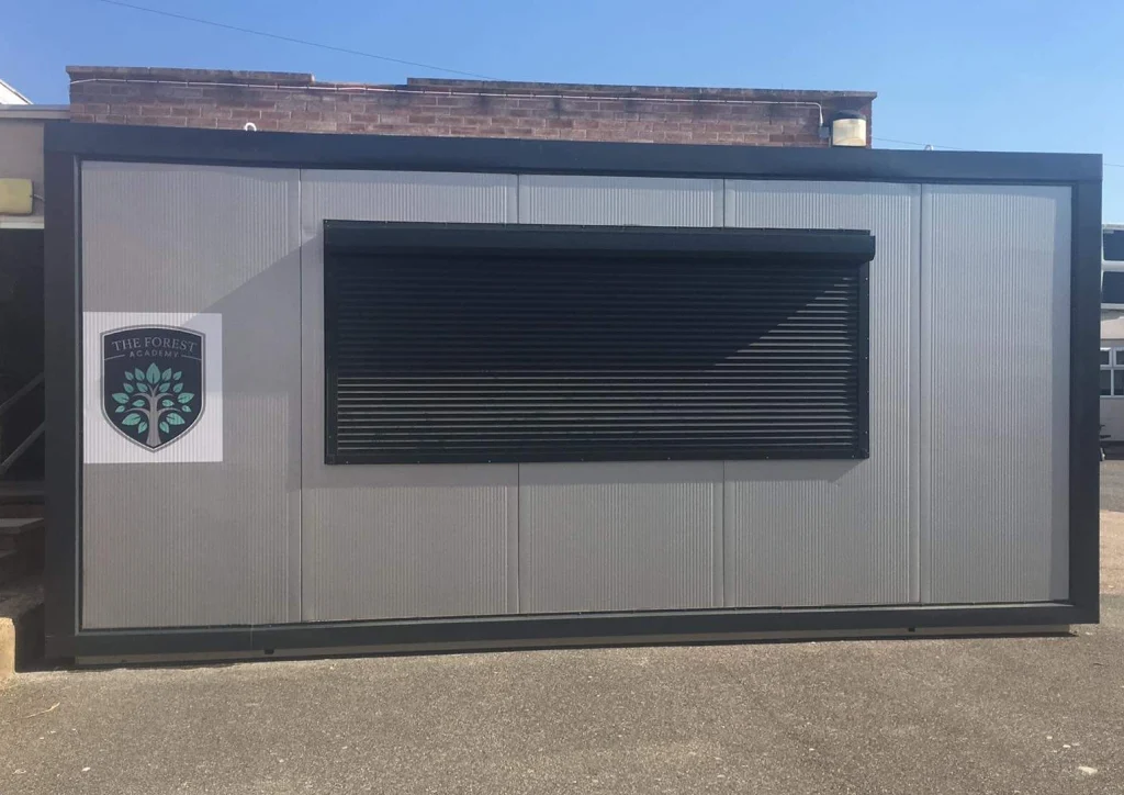 Dark modular kiosk or bar unit with timber panel cladding and roller shutter window in winter yard