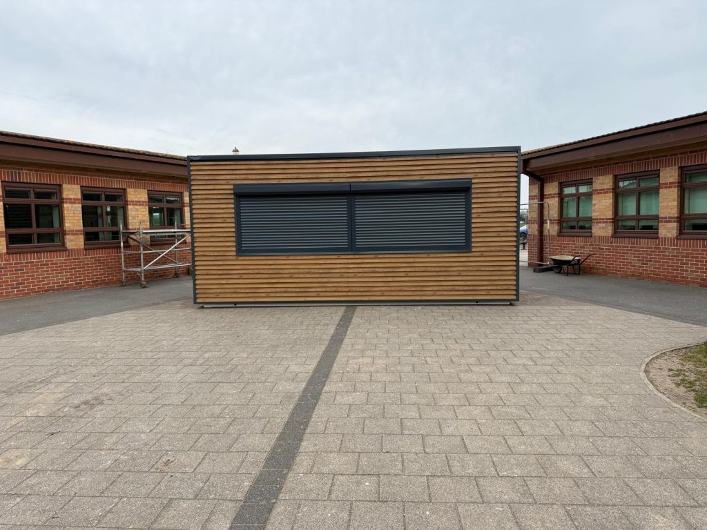 Timber-clad portable kiosk with dual roller shutters installed in a school playground between brick buildings
