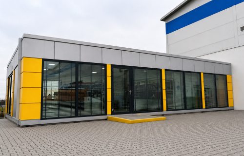 Exterior of a grey modular office building with yellow accent panels and large glazed frontage on a paved forecourt