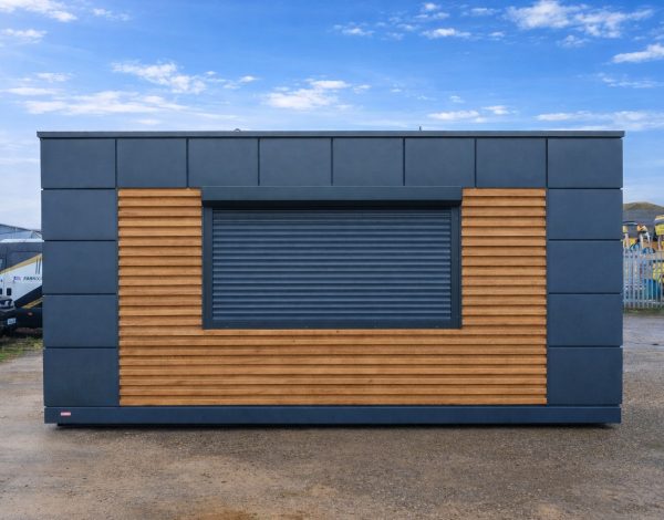 Portable modular kiosk with dark grey cladding, timber slat panels, and closed roller shutter hatch on site