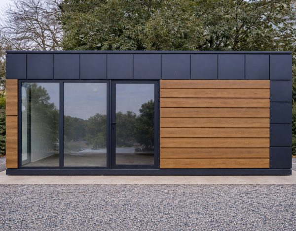 Front elevation of dark modular building with horizontal timber cladding and wide sliding aluminium-framed glass doors on gravel pad
