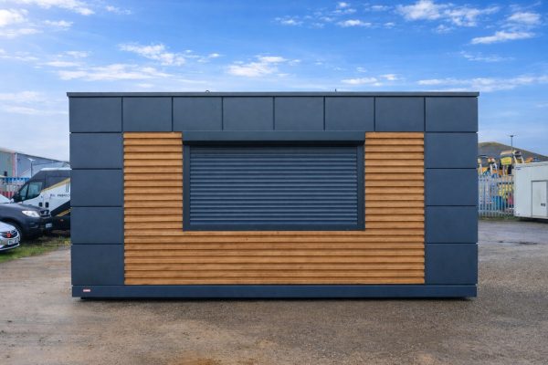 Portable modular kiosk with dark grey cladding, timber slat panels, and closed roller shutter hatch on site