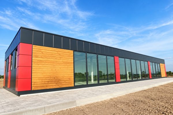 Large modular building with dark grey anthracite panels, red accent cladding, and wide timber section with full-length glazing