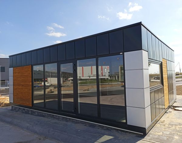 Modular building with floor-to-ceiling glazed facade dark grey cladding and wood accent panel on paved site